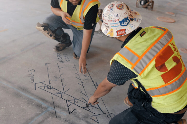 Two construction workers in safety vests and hard hats reviewing and marking conduit layout plans directly on a concrete floor as part of Lean construction planning.